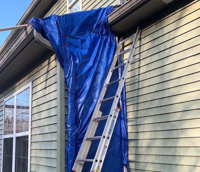 Roof with blue tarp and ladder against side of home